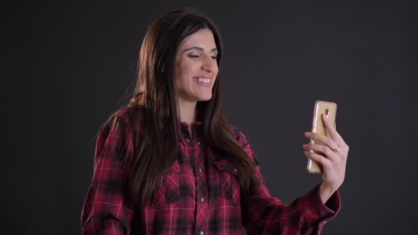 Portrait de profil de jeune et belle fille caucasienne aux cheveux longs parlant en vidéochat à l'aide d'un smartphone sur fond noir . — Video