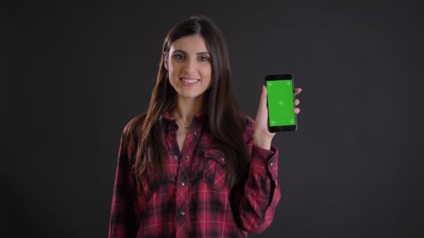 Retrato de una joven caucásica de pelo largo que sonríe mostrando la pantalla verde del teléfono inteligente sobre fondo negro . — Vídeo de stock