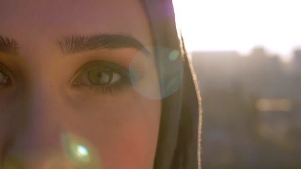 Retrato de primer plano de media cara de una joven mujer caucásica en hijab mirando directamente a la cámara con la ciudad urbana en el fondo — Vídeos de Stock