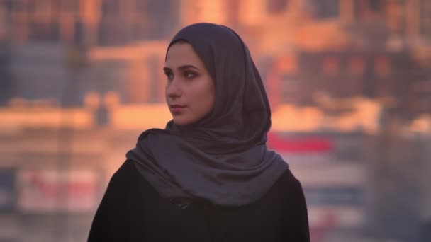 Крупный план портрета молодой привлекательной женщины в хиджабе, уверенно смотрящей в камеру с вечерним городом на заднем плане — стоковое видео
