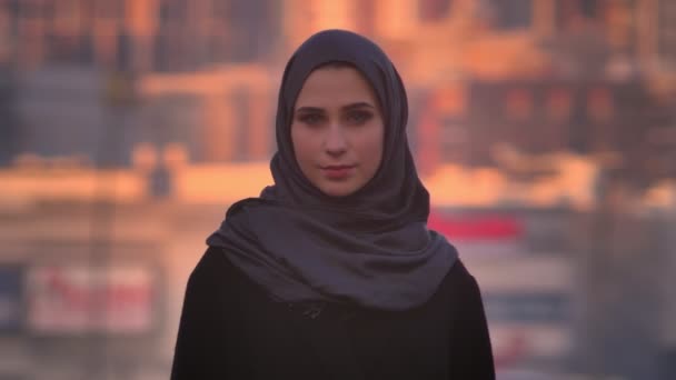 Primer plano retrato de joven atractiva hembra en hijab mirando directamente a la cámara con confianza con la ciudad en el fondo — Vídeos de Stock