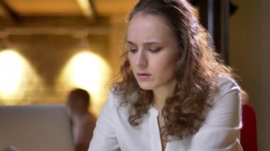 Yakın çekim dikkatle laptop ile çalışma ve ofiste içecek içme genç kıvırcık saçlı kadın portresi.