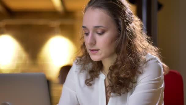 Gros plan portrait d'une jeune femme aux cheveux bouclés travaillant avec attention avec un ordinateur portable et buvant volontiers des boissons au bureau . — Video