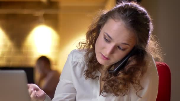 Close-up portret van curly-haired vrouw praten over cellphone en kijken in laptop gelijktijdig in kantoor. — Stockvideo
