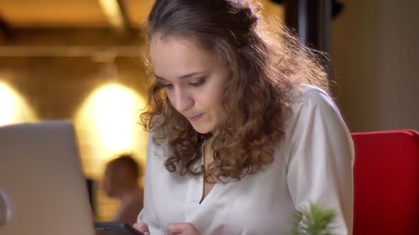 Portrait en gros plan d'une jeune femme d'affaires aux cheveux bouclés tapant judicieusement sur son smartphone au bureau . — Video