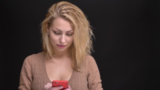 Ritratto di splendida donna caucasica dai capelli lunghi che sorride guardando nello smartphone su sfondo nero . — Video Stock