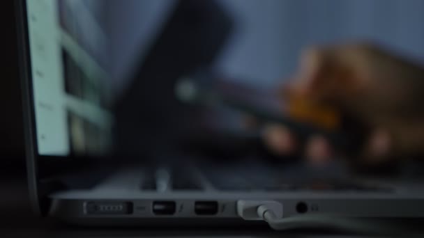 Tiro de close-up desfocado de mãos trabalhando com smartphone na frente do laptop no fundo escuro . — Vídeo de Stock