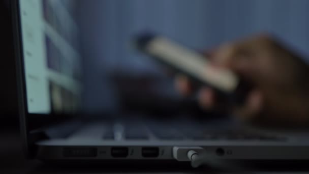 Θολή κοντινή λήψη των χεριών πληκτρολόγηση σε smartphone μπροστά από το φορητό υπολογιστή σε σκούρο φόντο. — Αρχείο Βίντεο