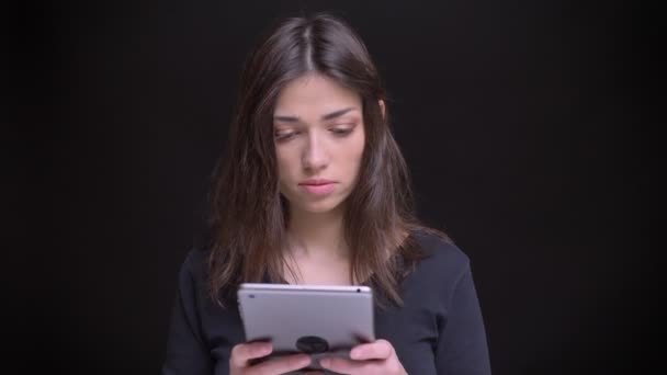 Retrato de bela menina morena de cabelos longos caucasiana atentamente assistindo em tablet no fundo preto . — Vídeo de Stock