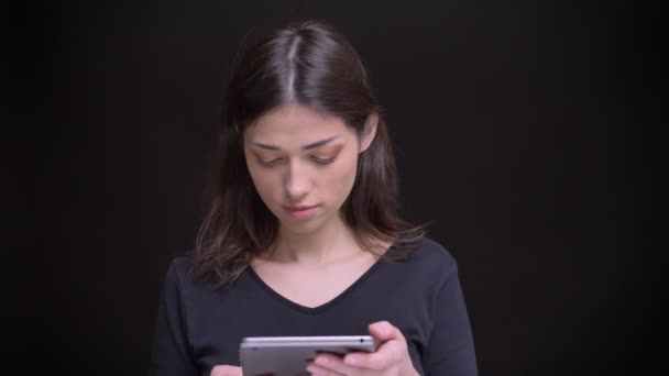 Retrato de chica morena caucásica de pelo largo mirando atentamente en la tableta sonríe en la cámara sobre fondo negro . — Vídeo de stock