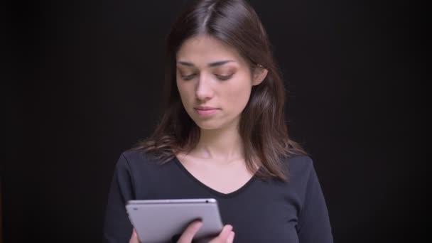Pretty long-haired brunette girl watching into tablet and smilingly shows its upright green screen into camera on black background. — Stock Video