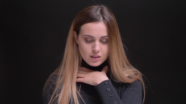 Portrait of young caucasian long-haired blonde girl having sore throat and watching sadly into camera on black background. — Stock Video