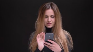 Genç beyaz uzun saçlı sarışın kız siyah arka plan üzerine smartphone içine dikkatle izlerken portresi.
