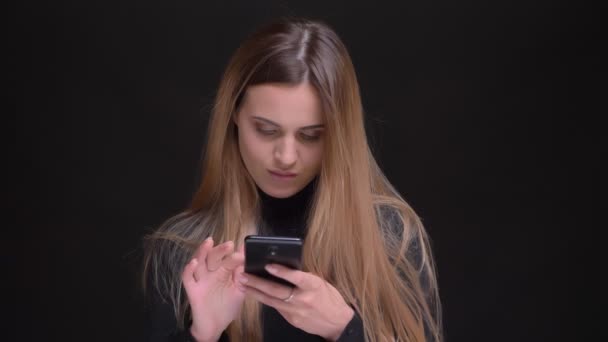 Porträtt av unga kaukasiska långhåriga blonda flicka glatt titta in smartphone på svart bakgrund. — Stockvideo