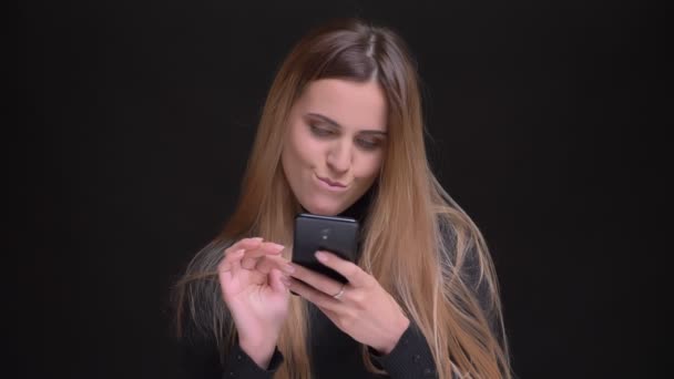 Portrait of young caucasian long-haired blonde girl joyfully watching into smartphone and biting lips on black background. — Stock Video