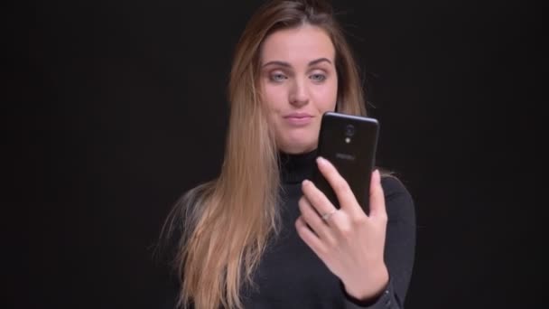 Retrato de una joven rubia caucásica de pelo largo haciendo fotos selfies alegremente usando un teléfono inteligente sobre fondo negro . — Vídeos de Stock