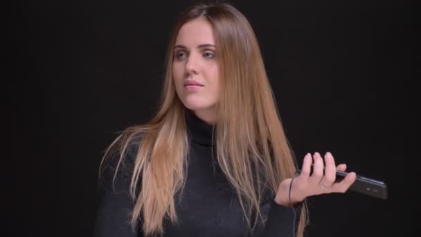Porträtt av unga kaukasiska långhåriga blonda flicka Lyssna på musik med smartphone och hörlurar på svart bakgrund. — Stockvideo