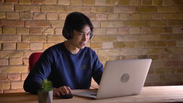 Retrato de close-up de jovem empresário coreano em fones de ouvido tendo uma chamada de vídeo no laptop dentro de casa — Vídeo de Stock