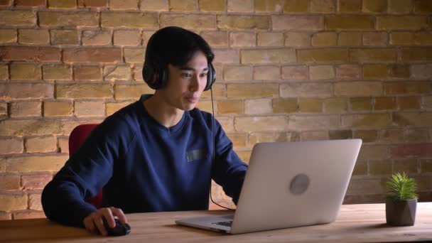 Retrato de close-up do jovem estudante coreano em fones de ouvido usando o laptop e ficando animado levantando braços — Vídeo de Stock