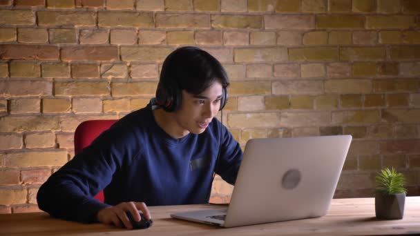 Retrato de close-up de jovem atraente empresário coreano em fones de ouvido usando laptop e rindo com uma emoção — Vídeo de Stock