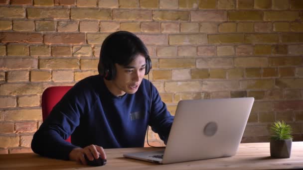 Closeup portret van jonge aantrekkelijke Koreaanse mannelijke student hoofdtelefoon controleren op de laptop en steeds boos en gefrustreerd. — Stockvideo