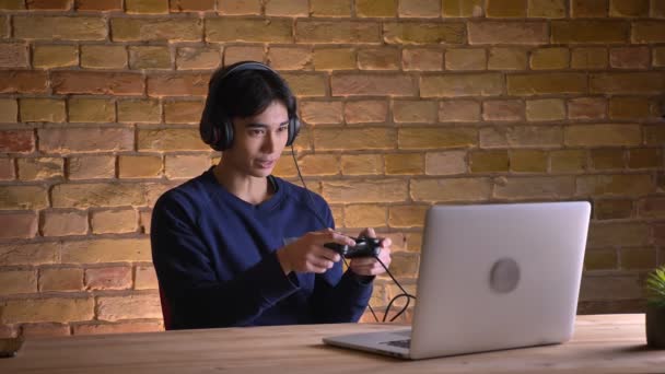 Retrato de primer plano del joven atractivo coreano en auriculares jugando videojuegos en la consola usando el portátil — Vídeos de Stock