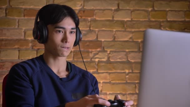 Primer plano retrato de joven atractivo hombre coreano en auriculares jugando videojuegos en interiores — Vídeo de stock