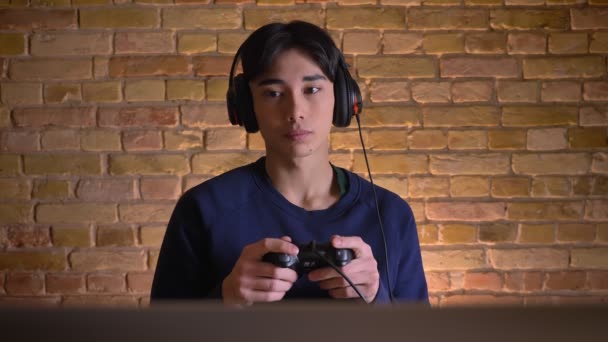 Крупный план портрета молодого привлекательного корейца в наушниках, играющего в видеоигры и выигрывающего — стоковое видео