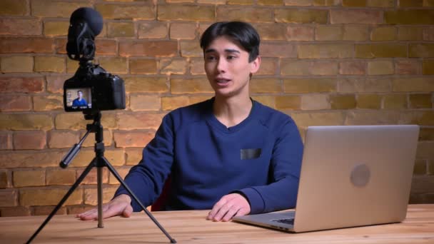 Retrato de cerca del joven blogger coreano hablando en cámara y saludando con su mano — Vídeos de Stock