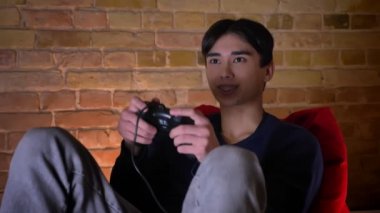 Portre portre kapalı fasulye çanta üzerinde otururken heyecanlı genç Koreli erkek video oyunları oynarken ve almak