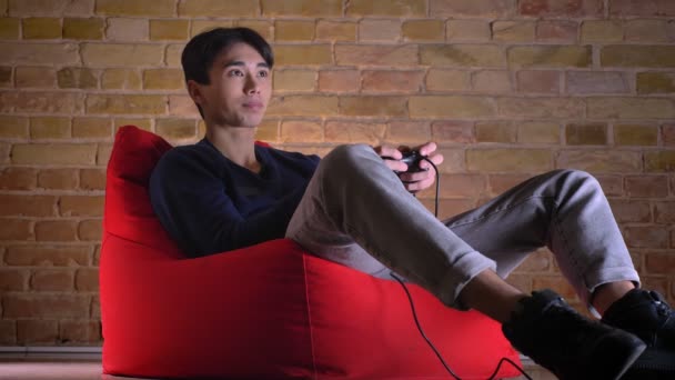Retrato de primer plano de un joven coreano jugando videojuegos que se pone feliz y sonríe mientras está sentado en la bolsa de frijoles — Vídeo de stock