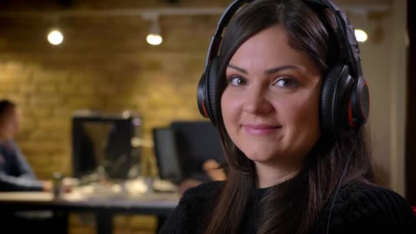 Retrato de close-up de empresária caucasiana adulta em fones de ouvido girando e olhando para a câmera sorrindo estar no local de trabalho — Vídeo de Stock