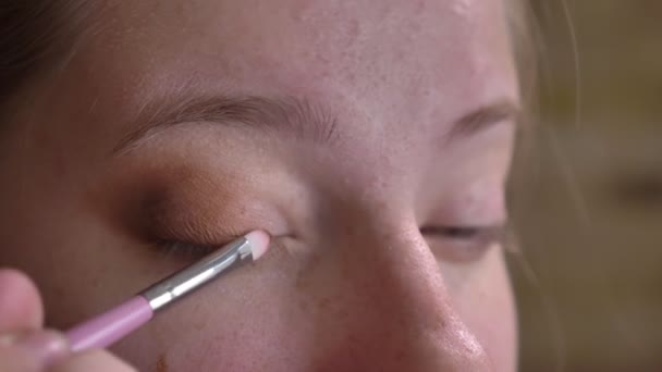 Retrato de primer plano de una joven artista de maquillaje caucásica que aplica sombra de ojos marrón en su párpado superior — Vídeos de Stock