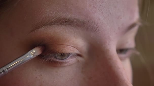 Retrato de primer plano de una joven artista de maquillaje caucásica que aplica sombras de ojos de color marrón oscuro en su párpado superior — Vídeos de Stock