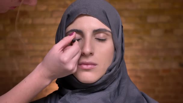 Manos femeninas haciendo maquillaje de ojos usando pensil negro y cepillo pequeño para mujer musulmana en hijab sobre fondo de pared de ladrillo . — Vídeo de stock