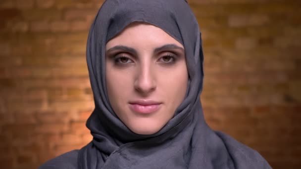 Portrait de femme musulmane en hijab avec maquillage des yeux fumés regardant calmement dans la caméra sur fond de mur de poulet . — Video