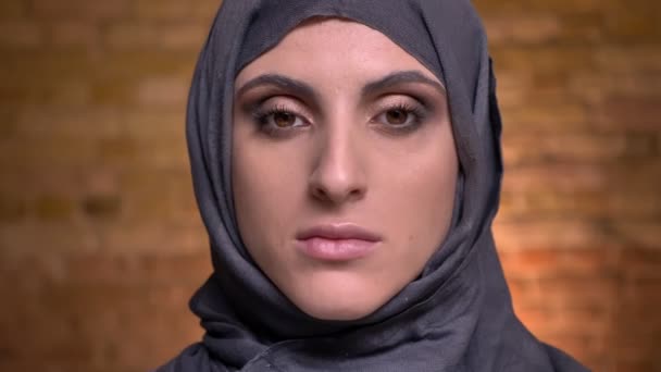 Porträtt av vackra muslimsk kvinna i hijab med ljusa make-up tittar allvarligt in kameran på bricken vägg bakgrund. — Stockvideo