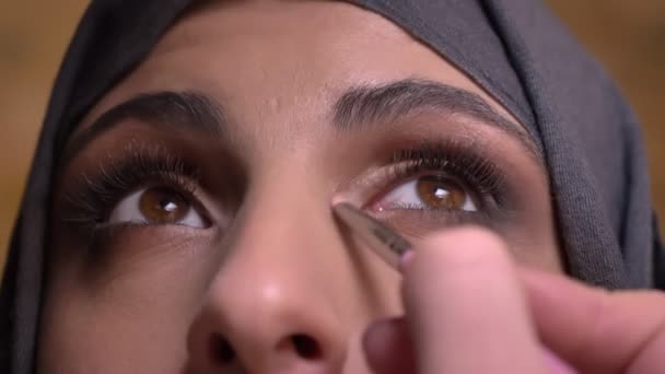 Портрет рук, делающих макияж глаз с небольшой кисточкой для мусульманки в хиджабе, смотрящей вверх на фоне кирпичной стены . — стоковое видео