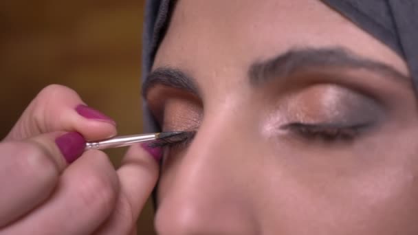 Portrait of female hands drawing black arrow on eye of beautiful muslim lady in hijab on bricken wall background. — Stock Video