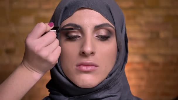 Portrait of female hands brushing eyebrows of muslim woman in hijab on bricken wall background. — Stock Video