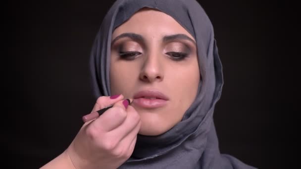 Retrato de manos femeninas dibujando labios usando pensil marrón para mujer musulmana en hijab sobre fondo negro . — Vídeos de Stock