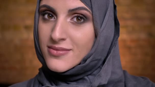 Retrato de una hermosa mujer musulmana en hijab con maquillaje brillante gira la cabeza y sonríe en la cámara sobre fondo de pared de ladrillo . — Vídeo de stock