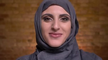 Hijab parlak makyajlı utanarak başını kapatarak ve içine bricken duvar arka plan kameraya gülümseyen güzel Müslüman kadın portresi.