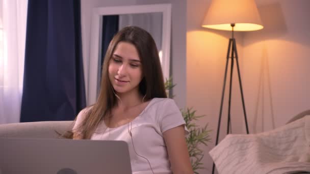 Retrato de cerca de una joven adolescente atractiva escuchando música en los auriculares usando el portátil en el dormitorio — Vídeo de stock