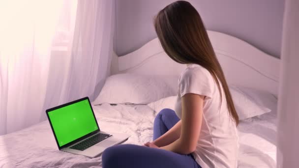 Retrato de cerca de una joven atractiva hembra caucásica mirando la pantalla verde de croma-key del portátil sentado en la cama y arreglando su cabello — Vídeos de Stock