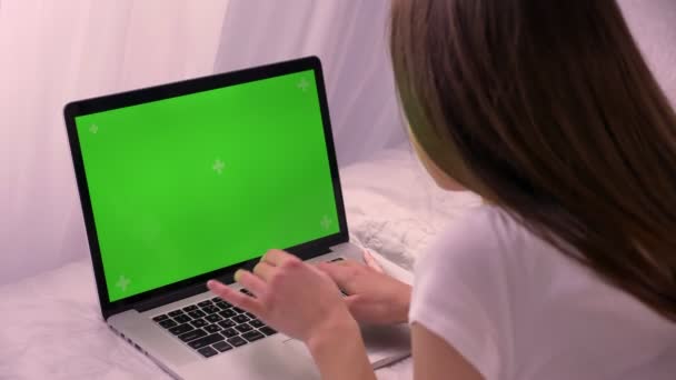Вид сзади на молодую привлекательную кавказскую женщину, печатающую на ноутбуке с зеленым цветным экраном, лежащим на кровати в помещении — стоковое видео