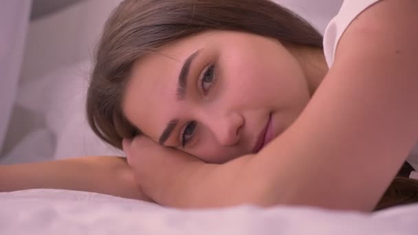 Closeup portrait of young stunning caucasian female lying on the bed looking at camera and daydreaming — Stock Video