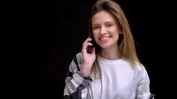 Retrato de jovem caucasiano de cabelos compridos menina em camisa xadrez sorrindo falando no celular e assistindo em câmera no fundo preto . — Fotografia de Stock