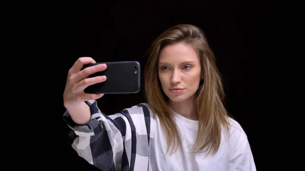 Portrait of beautiful young caucasian long-haired girl in shirt making selfie-photos using smartphone on black background. — Stock Photo, Image