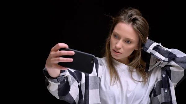 Портрет молодої кавказької довгошерстої дівчини в плетеній сорочці, яка торкається її волосся і робить селфі-фотографії, використовуючи смартфон на чорному тлі . — стокове фото